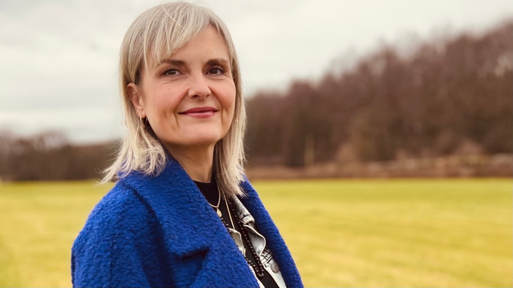 Linz Anderson, Somatic Renewal Coach, standing in woodland wearing a blue coat
